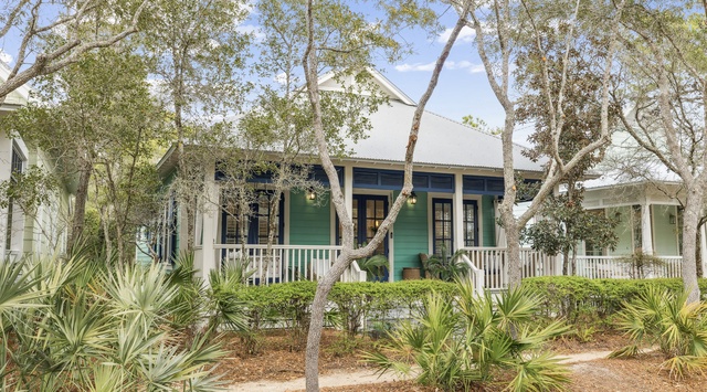 Charming green cottage nestled among mature trees and tropical landscaping, featuring a welcoming front porch and traditional coastal architecture.