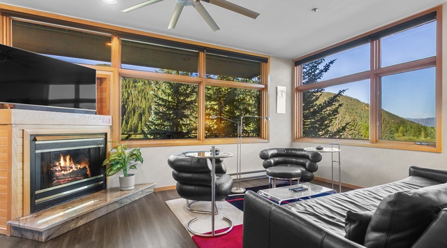 Unwind in this inviting living room where mountain views frame your relaxation. Sink into modern seating by the cozy fireplace, enjoying peaceful forest scenery through expansive windows.