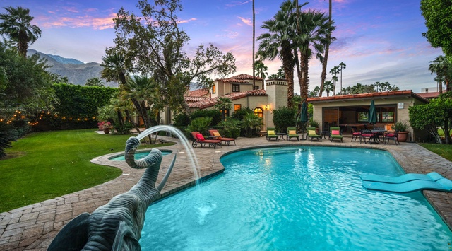 A stunning desert oasis awaits with sparkling pool, lush lawn, and mountain views framed by towering palms. This Spanish-style villa offers your perfect Palm Springs escape at sunset.