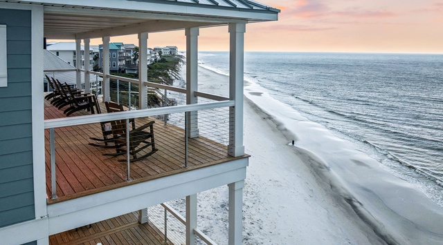 The property features two private balconies with sweeping beachfront views. Each level offers rocking chairs overlooking white sand and gentle surf, framed by a stunning coastal sunset.