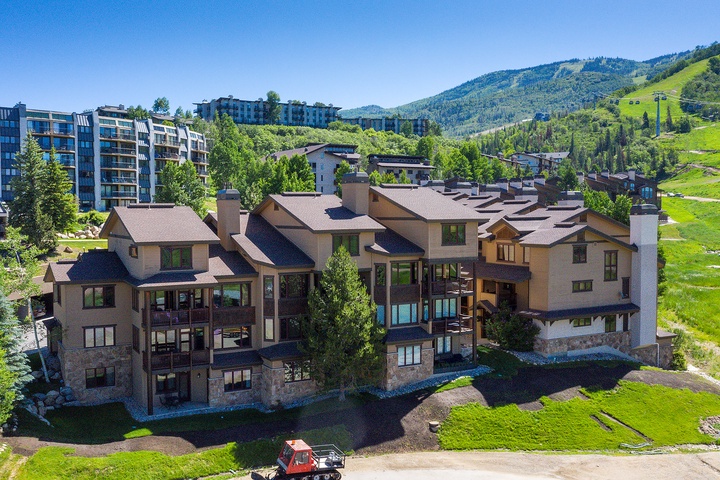 Multi-level mountain resort buildings nestled in a scenic valley setting, surrounded by lush greenery and rolling hills with ski slopes visible in the distance.