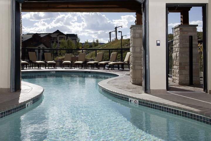 A winding outdoor pool surrounded by lounge chairs offers a peaceful setting with mountain views and neighboring homes, accessed through a shaded stone-framed entrance.
