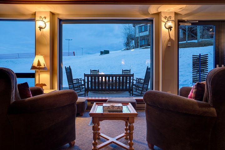 Curl up in plush seating as you gaze through expansive windows framing a serene winter wonderland, where gentle lamplight creates a warm, inviting glow against the snowy landscape outside.