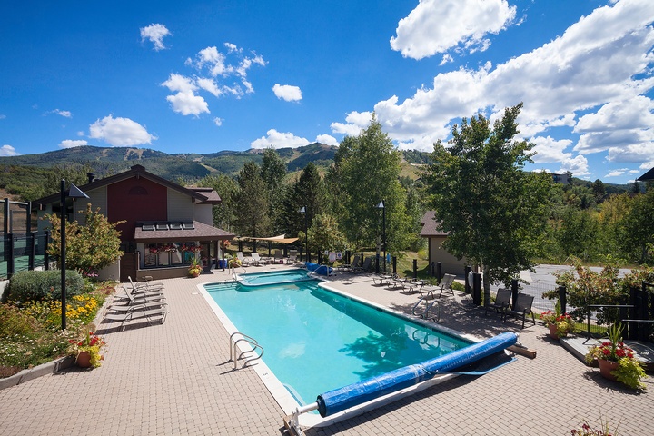 A scenic mountain resort features an inviting outdoor pool surrounded by lounge chairs, set against lush greenery and dramatic mountain peaks under a clear blue sky with scattered clouds.