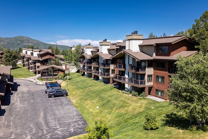 A mountain resort community featuring multi-level alpine-style condominiums with private balconies, expansive lawns, parking, and forested peaks beyond.