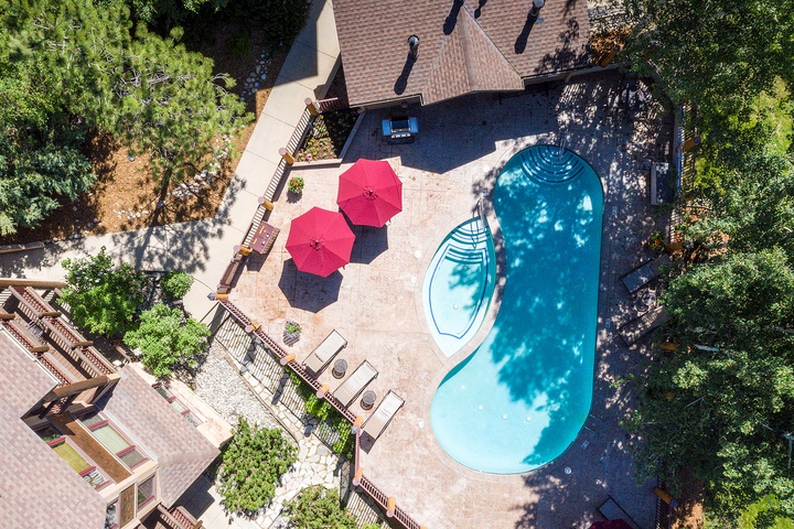 Aerial view of the property showcasing a kidney-shaped swimming pool, hot tub, patio with red umbrellas, and lush surrounding greenery.