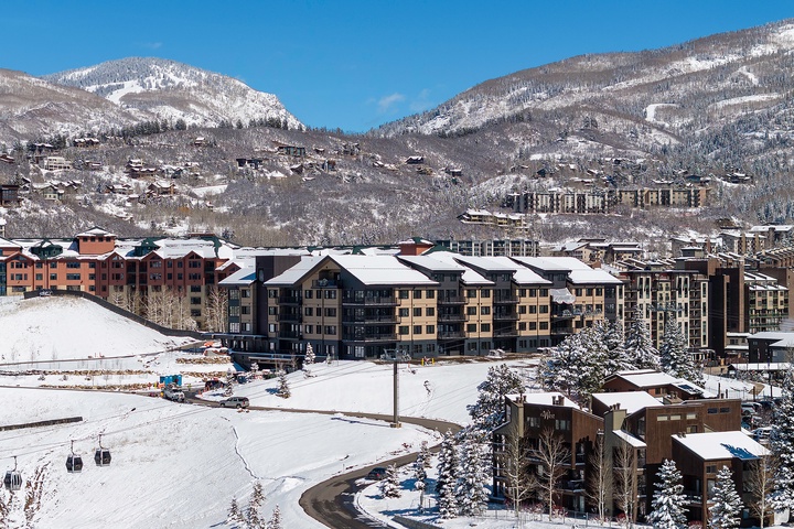 A snow-covered mountain resort village nestled in a valley, featuring multi-story lodges, ski lifts, and scenic alpine peaks under crisp blue skies.