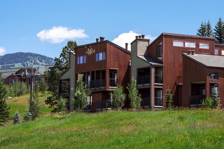 Alpine vacation property exterior featuring multi-level wood and siding architecture nestled against green ski slopes and mountain scenery under clear blue skies.