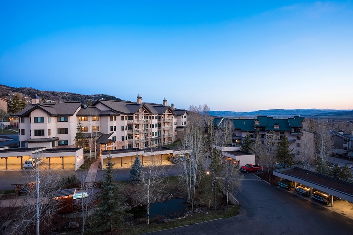 A mountain resort community nestled in the valley, featuring multi-level accommodations with covered parking, surrounded by evergreens and framed by distant peaks under an open sky.