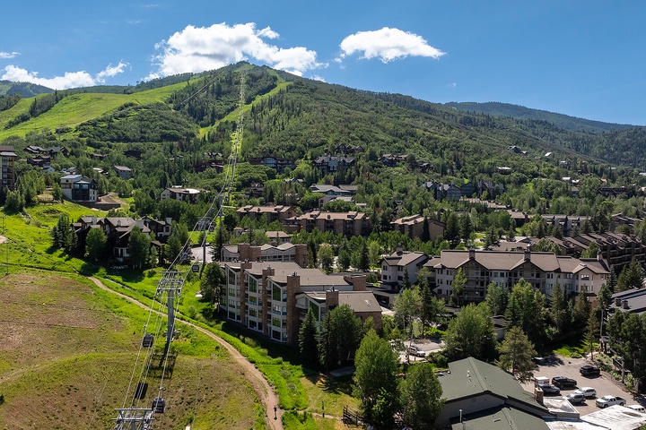 Aerial view of a mountain resort community featuring ski slopes, residential buildings, and forested hillsides during summer season with clear blue skies.