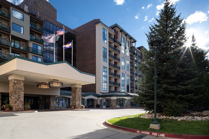 Modern multi-story property featuring stone-accented entrance, manicured grounds, and welcoming covered drive. Located in a peaceful mountain setting with mature evergreens and blue skies overhead.