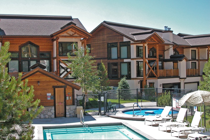 This mountain resort property features multi-story timber and stucco buildings with alpine-style architecture, surrounded by evergreen trees and equipped with outdoor pools, hot tubs, and a sauna house.
