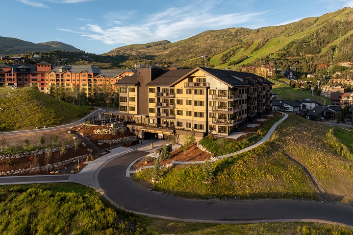 This aerial perspective showcases the property nestled in a picturesque mountain valley, surrounded by lush summer greenery and sweeping hillside vistas. The building is part of a scenic resort community with easy access to outdoor recreation.