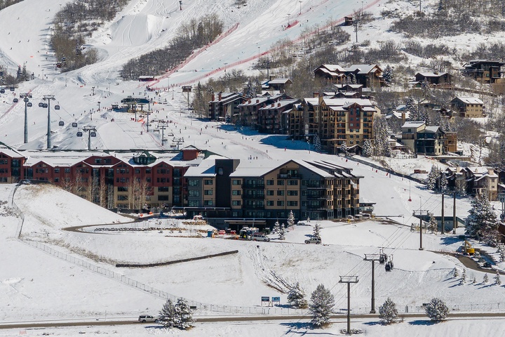 Aerial view of a mountain ski resort village nestled at the base of snow-covered slopes, featuring ski lifts and multiple lodging buildings in a winter alpine setting.