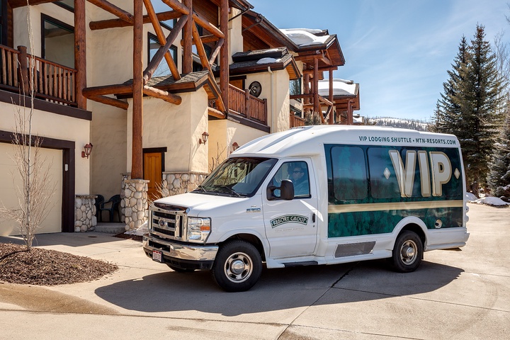 A VIP lodging shuttle van parked in front of a mountain resort property with timber-frame architecture and snow-capped peaks in the background.