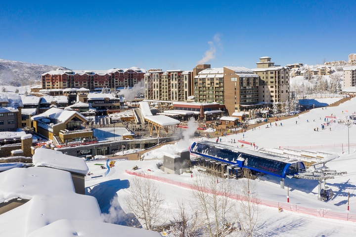 A vibrant ski resort village with snow-covered slopes, modern lodge buildings, and mountain gondolas set against a backdrop of snow-capped peaks under clear blue skies.