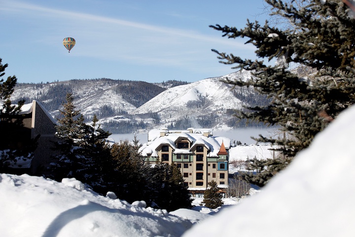A charming alpine property nestled against snow-covered mountain peaks, framed by evergreen trees, with a hot air balloon drifting overhead in a crisp winter sky.