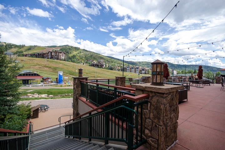 A mountain resort setting with expansive alpine views, featuring outdoor terraces and hillside accommodations surrounded by forested peaks and summer skies.