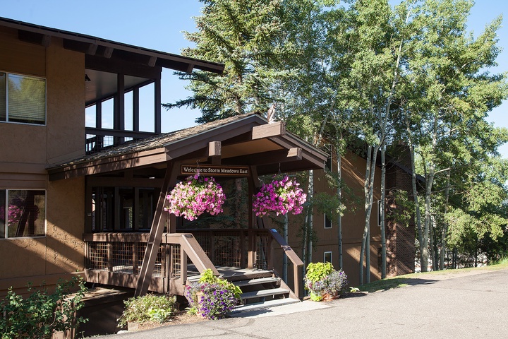 Storm Meadows Base welcomes guests with a charming covered entrance adorned with vibrant hanging flower baskets, surrounded by towering trees and mountain scenery.