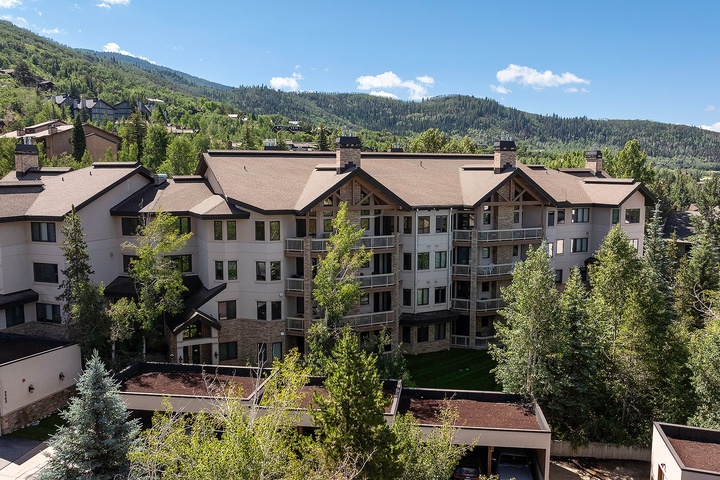 Multi-story mountain lodge nestled in lush evergreen forests with expansive views of tree-covered peaks under clear blue skies.