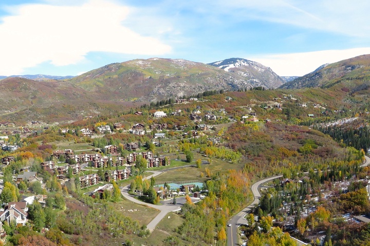 Aerial view of a mountain resort nestled in a scenic valley with autumn foliage, residential buildings, and winding roads framed by forested peaks.