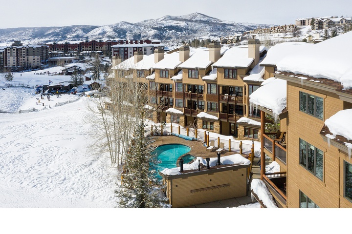 A mountain resort property featuring snow-covered buildings with an outdoor hot tub, nestled among snowy slopes and surrounded by ski resort amenities and mountain scenery.