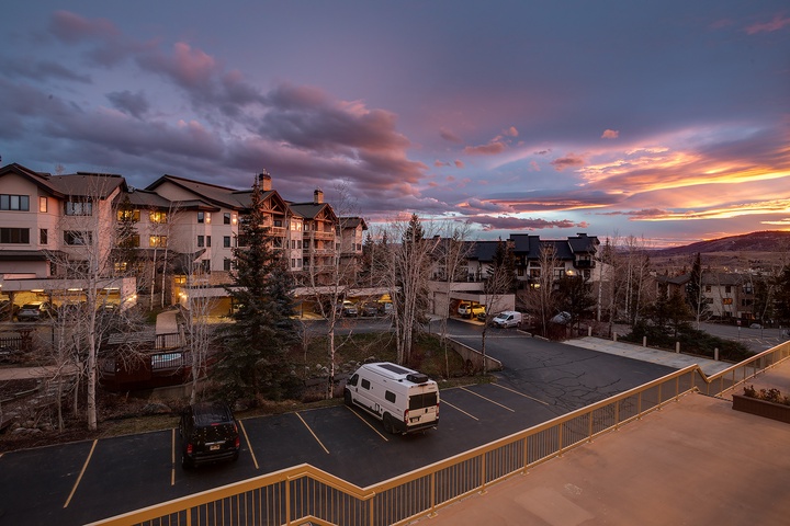 A dramatic sunset paints the sky in vibrant hues above this mountain resort property, with multiple buildings nestled among evergreens and barren winter trees in the surrounding area.
