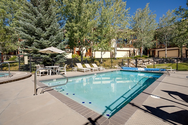 Communal pool area set in a peaceful, tree-lined neighborhood. Features sunny deck space with lounge chairs, umbrella seating, and mountain-style buildings in the background.