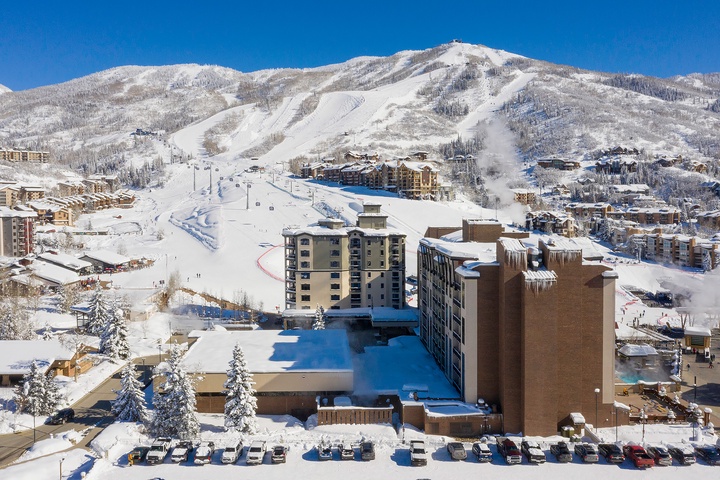Aerial view of a premier ski resort nestled at the base of snow-covered mountain slopes, with ski lifts and trails winding through frosted evergreens under a brilliant blue winter sky.