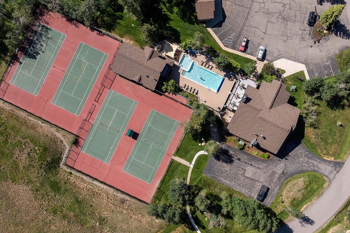 Aerial view of the property showcasing tennis courts, a sparkling pool, and ample parking surrounded by lush greenery and natural landscape.