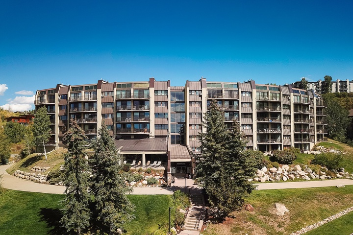 A multi-story mountain resort building with spacious balconies, nestled in a scenic alpine setting with manicured lawns, evergreen trees, and natural stone landscaping.