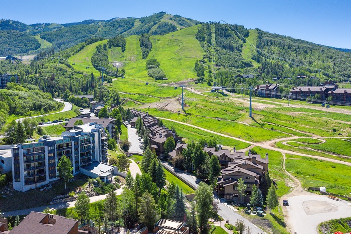 Aerial view of a mountain resort community nestled at the base of summer ski slopes, surrounded by lush green meadows and forested peaks.