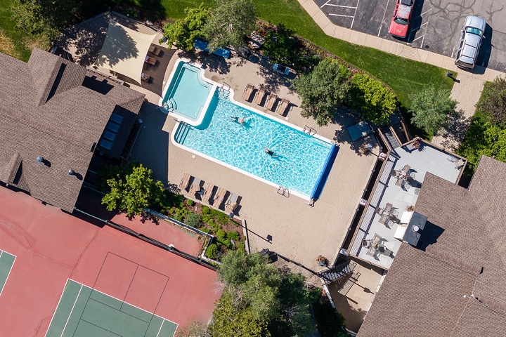Aerial view of the property featuring a sparkling outdoor swimming pool surrounded by lounge chairs, tennis court, and landscaped green spaces with ample parking nearby.