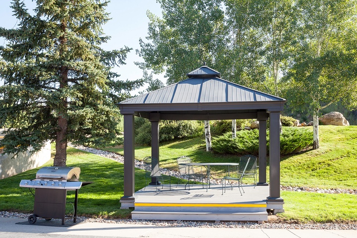 A covered pavilion with dining seating and a BBQ grill nestled in landscaped grounds, perfect for relaxing outdoor meals surrounded by evergreens and rolling lawns.