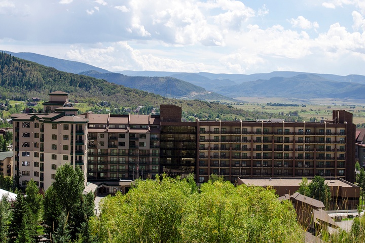 Multi-story resort building set against scenic mountain terrain with forested hillsides and a valley floor visible in the background, surrounded by trees and offering elevated views of the landscape.