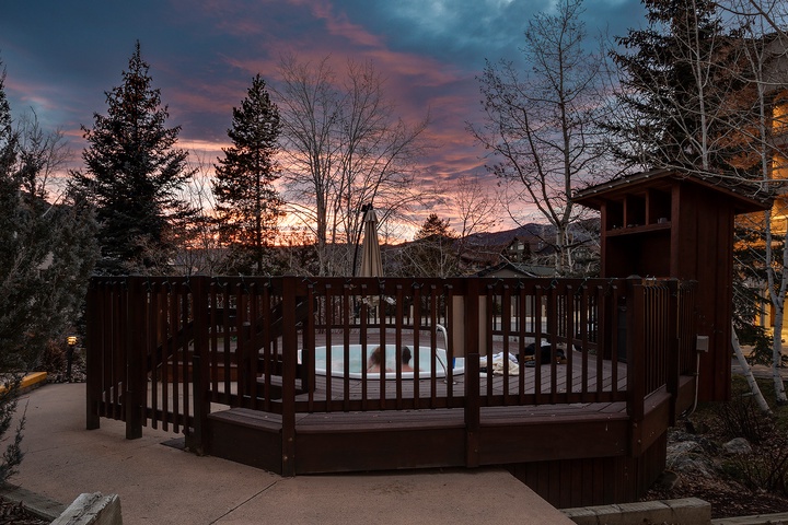 Property features a hot tub area surrounded by wooden railings, set against a backdrop of evergreen trees and colorful twilight sky.
