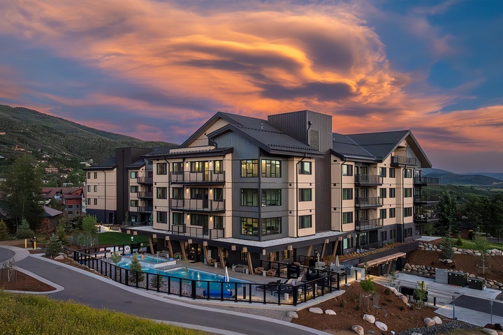 A modern mountain resort property showcasing contemporary architecture with dramatic cloud formations overhead, nestled among rolling hillsides and neighboring structures in the surrounding valley.