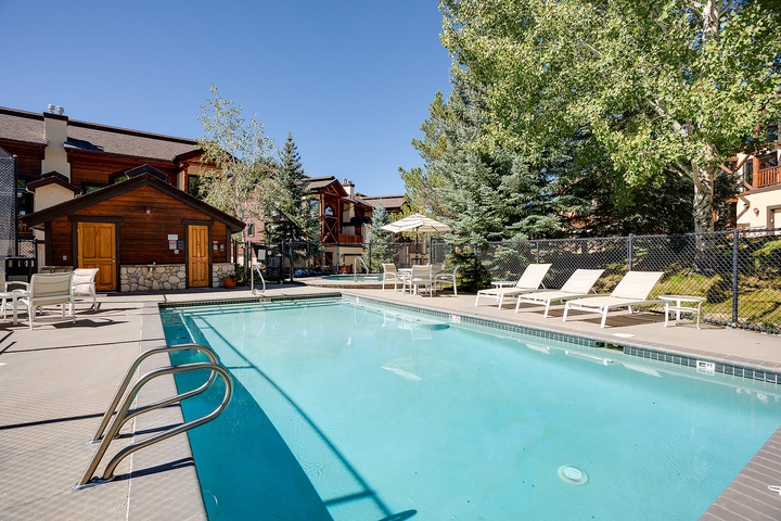 Outdoor swimming pool surrounded by mature trees and wooden cabins, with loungers and patio umbrella for guest relaxation during warmer months.