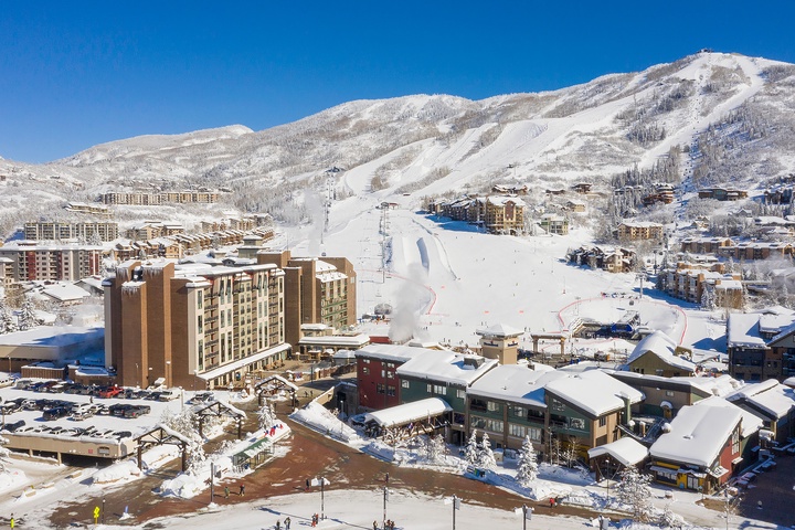 Aerial view of a premier ski resort village nestled in snow-covered mountains, featuring ski-in/ski-out access, ski lifts, and alpine lodges under clear blue skies.