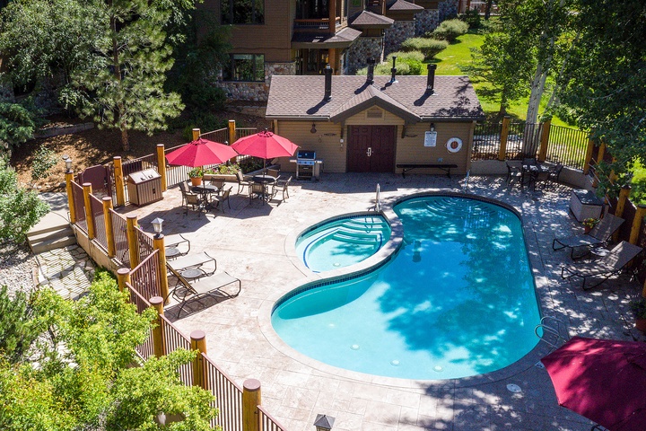 Aerial view of the property featuring a private pool, hot tub, and patio dining area with BBQ facilities, surrounded by lush trees and mountain greenery.