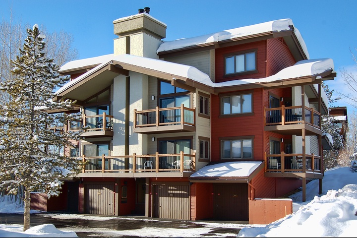 Modern ski chalet nestled in snowy mountain landscape, featuring warm red cedar siding, multiple balconies, and inviting covered entry with chimney rising above snow-capped roofline.
