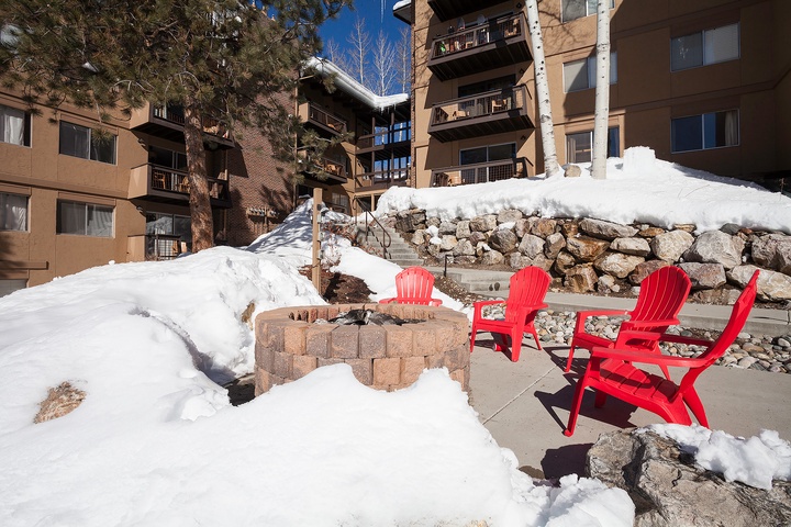The property sits in a winter mountain setting with bright red seating around an outdoor fire pit, snow-covered stone walls, and multi-story buildings nearby.