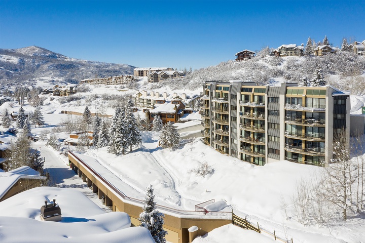 This alpine village resort nestled in snow-covered mountains offers stunning winter scenery. Modern multi-story buildings sit amid snowy pines, with ski lodges dotting the mountainside under clear blue skies.
