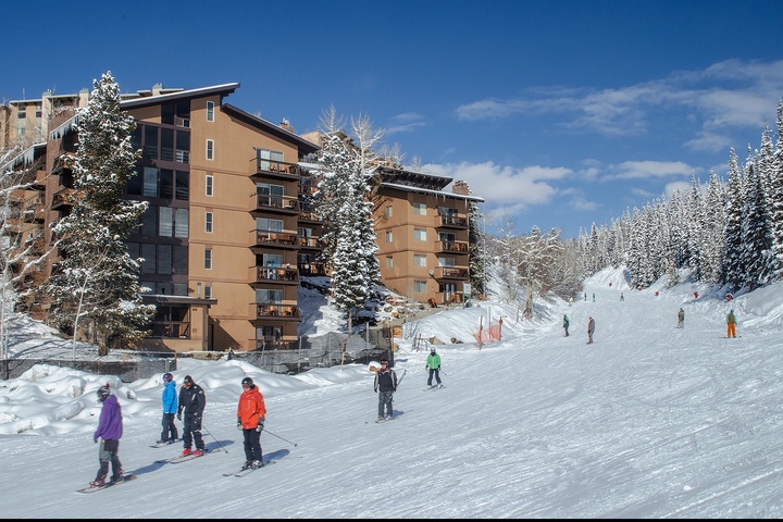 Multi-story mountain resort buildings nestled at the base of ski slopes, offering convenient ski-in/ski-out access surrounded by snow-covered evergreens under blue skies.