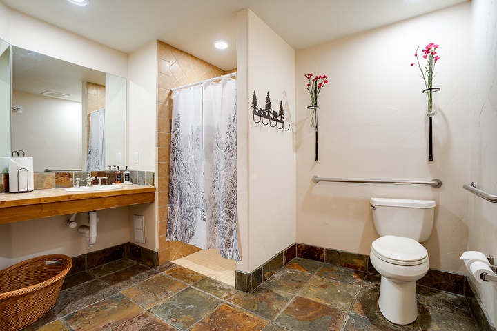 Your spacious bathroom features a walk-in shower with forest-themed curtain, warm tile flooring, and thoughtful grab bars for comfort and safety. Fresh flowers add a welcoming touch to your mountain retreat.