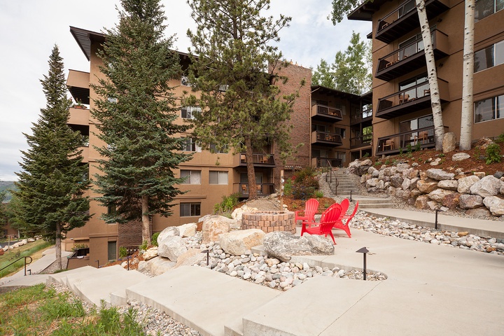 The property features multi-story residential buildings nestled among evergreens, with landscaped outdoor spaces including a fire pit and red Adirondack chairs for guests to enjoy the mountain surroundings.