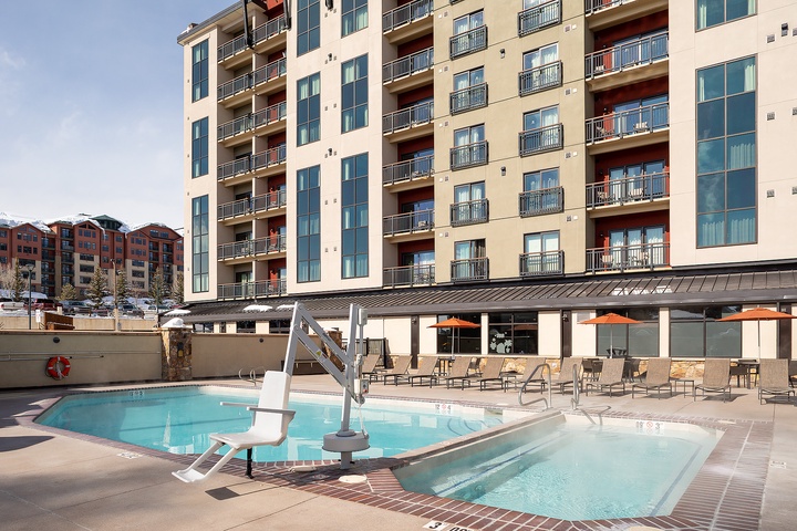 The property features a modern multi-level design with private balconies and an outdoor pool area complete with lounge seating and covered dining space, set against a scenic mountain backdrop.