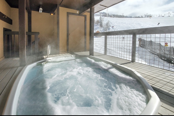 A bubbling hot tub on a covered terrace offers stunning views of snow-covered ski slopes and mountain scenery, perfect for relaxing après-ski moments.
