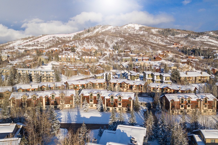 Winter scene featuring snow-covered vacation rentals nestled among evergreen trees at the base of a mountain, with ski slopes visible on the hillside.