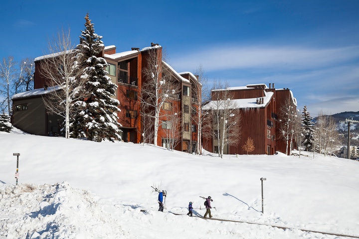 A multi-story mountain lodge complex sits nestled among snow-covered evergreens and aspens, with ski-in access visible on the snow-covered slopes below.
